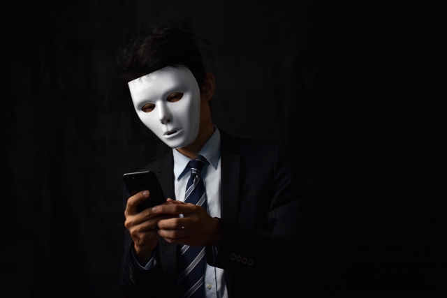 仮面をつけてスマホを操作する匿名ユーザー。怒りのままに発信する危うさを象徴